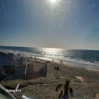 The image shows a beachfront scene, possibly in the Al-Mawasi area of an unknown city. The perspective is unusual; likely taken from an elevated or angled position. The beach has a sandy texture with scattered vegetation and makeshift structures, possibly shelters or market stalls. Several people are visible along the shoreline. The sea has a bright, reflective surface due to the sun, positioned in the upper-left of the frame. The sky is mostly clear, light blue with some haziness near the horizon. The words "Galaxy A14" are visible in the lower-right corner, indicating the device used to take the photo.