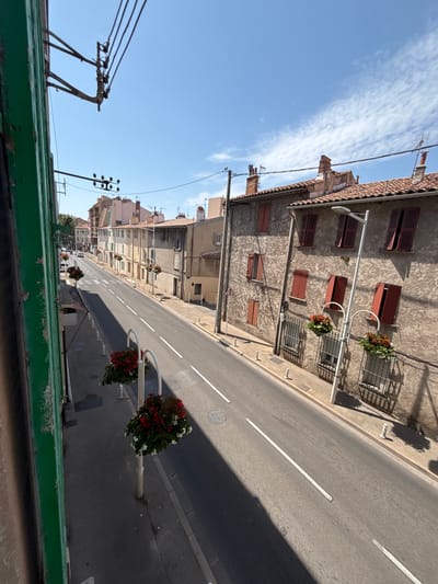 This is a high-angle, full shot of a street in Toulon, France, on a clear, sunny day. The street is paved and marked with white lines. On the right side of the street, there are two- and three-story buildings with tiled roofs and red shutters. Many of the windows are shuttered. There are streetlights along the sidewalk with hanging baskets of red and white flowers. The sidewalk is concrete. On the left side of the street, there are more buildings, some of which are multi-colored. Utility poles and wires are visible. A few cars are driving down the street. The foreground is dominated by a green painted wall, likely the exterior of a building. The overall scene depicts a quiet, residential street in what appears to be a European city.