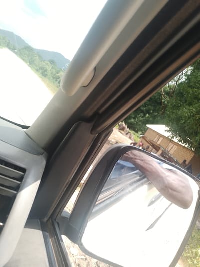 The image is taken from inside a vehicle in Serti, Nigeria, showing a view through the windshield and a side mirror. The exterior scene includes a dirt road, a partially constructed building with a corrugated metal roof, and lush green trees. The building appears to be made of mud bricks. Several people are visible outside the vehicle, some standing near the building. The side mirror reflects the arm of a person and the interior of the vehicle. The interior shows part of the dashboard, a vent, and the interior structure of the car's A-pillar. The sky is bright and appears to be daytime.