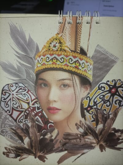 A photograph captures a close-up, slightly overhead view of a page from a spiral-bound book or calendar, resting on a dark, flat surface in an indoor setting. The page, made of light-colored, textured paper, is secured at the top by four white metal spiral binding rings.
The central subject on the page is a composite image or collage featuring the head and upper chest of a young adult female with fair skin, dark hair, and lightly made-up features, gazing directly at the viewer. She is adorned with an elaborate golden, red, and white patterned crown, characteristic of traditional Dayak headwear, which includes what appear to be small feathers or beaded elements. Surrounding her are stylized grey feathers on the left, and two shield-like or fan-like objects with distinct black, yellow, red, and white curvilinear patterns, typical of Dayak tribal art, positioned on either side of her face. Clustered brown, feather-like elements are arranged at the bottom of the composition, with faint grey patterns, possibly textile motifs, serving as background elements within the collage.
The immediate environment visible behind the page is dark and out of focus. The upper right background shows a portion of a glowing computer monitor, displaying faint text or graphical elements, with a small green indicator light visible on a device.
The image depicts traditional cultural elements associated with the Dayak people, indigenous to the island of Borneo (Kalimantan, Indonesia). The physical object (the spiral-bound page) and the scene photographed are located indoors within the Special Capital Region of Jakarta, Indonesia.
