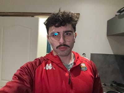 A man with glasses and a mustache wears a red Kappa jacket with an Italian Olympic emblem on the chest. He looks directly at the camera with a neutral expression. His hair is curly. The background is a plain white wall with a glimpse of a kitchen counter on the left. The image appears to be inverted.