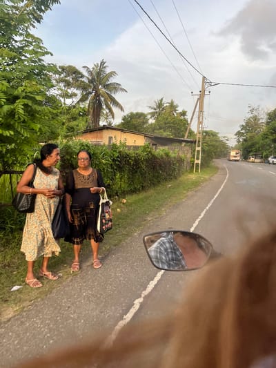 This media file, taken in Peleana, Sri Lanka, depicts an outdoor scene on a road. Two women are standing on the grassy shoulder of the road, facing each other. The woman closer to the foreground is wearing a light-colored, floral-patterned dress and a dark handbag. The woman behind her is wearing a dark dress with intricate gold and black detailing and also has a handbag. Both women are wearing sandals.
The setting appears to be rural, with lush green foliage and palm trees visible. In the background, a building with brick accents and a yellow facade can be seen. A utility pole with electrical wires runs along the side of the road. The road itself has a white dividing line. In the distance, a few vehicles, including what appears to be a bus, are driving on the road.
The time of day appears to be late afternoon or early evening, as the sky is overcast and the light is soft. The weather seems to be humid, typical of Sri Lanka. The overall mood is relaxed and casual, suggesting a chance encounter or a stop along a journey. A motorcycle's side mirror is visible in the foreground, reflecting the driver's checkered shirt, suggesting the photo was taken from a motorcycle.