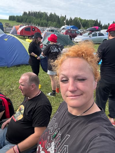 The image depicts an outdoor gathering in a grassy field in Большая Кивара, Russia, under an overcast sky, with a dense coniferous forest forming the distant background. In the foreground, a woman with curly red-orange hair, wearing a black t-shirt with a white graphic and a thin black necklace, faces the camera with a slight smile. To her left, a man with a receding hairline and a beard, wearing a black t-shirt with a yellow and red graphic, blue jeans, a pink wristband, and a dark red beaded necklace, is seated and looking towards the right. A red and black bag is positioned next to him.
In the mid-ground, multiple individuals are visible. A woman wearing a black bucket hat, black clothing, and a yellow wristband, holding a light-colored pouch, stands facing right, partially obscured by a large blue tent with yellow trim. Adjacent to her, another person, seen from the back, wears a red bucket hat, a black t-shirt with "MISFITS" written in white, white shorts, orange socks, and black and white sneakers. To the far right, partially obscured by the foreground woman's hair, a man in a red cap, black t-shirt, and black pants is visible from behind, wearing red flip-flops.
The field is populated with various camping tents, including orange, grey, and green structures. A prominent red teardrop-shaped camper trailer with a black "8" logo and "PANORAMA" text is centrally located. Numerous passenger vehicles, including a silver sedan with a black rooftop cargo box and a maroon SUV, are parked in the background. A wooden fence runs horizontally across the mid-distance, and a tall, thin pole extends upwards towards the sky in the far background.