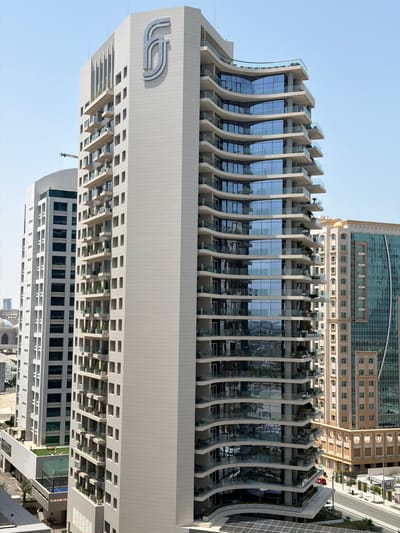 The image shows a close-up, angled view of a modern high-rise building with a curved facade on the right, featuring multiple balconies with glass railings and reflective windows. To its left, another taller, rectangular building with a lighter facade and smaller windows is visible, along with portions of other buildings and a crane in the background. The scene is set against a clear blue sky. The building on the right has a distinctive architectural design with undulating levels. The overall impression is of a contemporary urban landscape in a sunny climate. The logo "FS" is visible on the upper section of the building on the left. This building is part of the Lusail city development in Qatar.