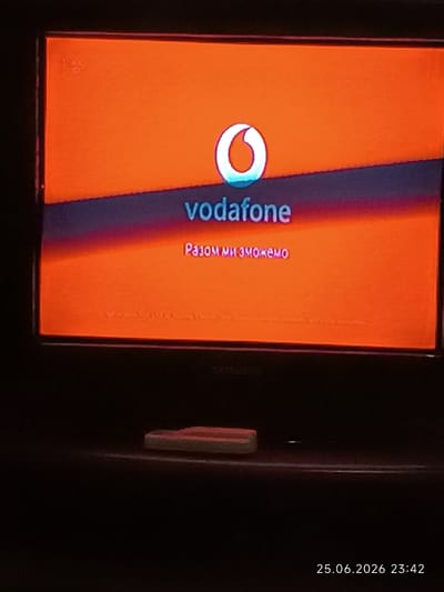 A television screen displays the Vodafone logo and text, with a date and time stamp at the bottom right. The screen is orange and purple, and the Vodafone logo is white. The text "vodafone" is written in white below the logo. Below that, in a pinkish-purple color, the text reads "Разом ми зможемо". In the bottom right corner, a timestamp reads "25.06.2026 23:42". The television appears to be in a dark room, with a remote control or similar object visible on the shelf below it. There is no indication of the city of Bolhrad, Ukraine being present in the image.