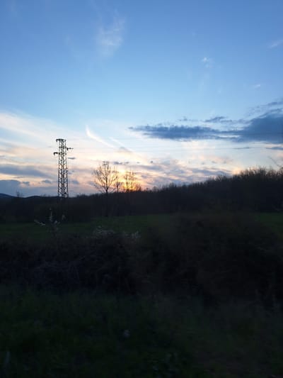 The image depicts an outdoor scene at dusk, likely in a rural or semi-rural area near Batoshevo, Bulgaria. A metal electricity pylon is silhouetted against a sky filled with light blue, white, and dusky pink clouds. Power lines stretch from the pylon into the distance. To the right of the pylon, there is a line of vegetation with trees and green grass. The vegetation is in varying degrees of light and shadow, possibly due to the setting sun. A bare tree stands silhouetted against the sky. The bottom of the image reveals the top of a hill. The sky is the most prominent feature, making up roughly half of the image.