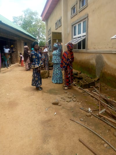 The image depicts an outdoor scene in Madakiya, Nigeria, likely a residential or community area. In the foreground, a sandy dirt path leads towards a building with a light-colored facade and a red-tiled roof. Several individuals, predominantly women wearing colorful traditional attire, are gathered. Some are standing and conversing, while others appear to be observing. One woman in the center is wearing a dark blue dress with circular patterns and a headscarf. To her right, another woman is dressed in a vibrant red and blue patterned outfit.
To the left of the main building, a smaller structure with a green corrugated metal roof is visible. A group of people are standing near the entrance of this structure. The ground is uneven, with scattered rocks, debris, and construction materials such as metal pipes and concrete blocks. Vegetation, including trees and grass, can be seen in the background, suggesting a semi-rural or transitional urban environment. The sky is overcast. The overall impression is that of a community gathering or an event taking place outdoors.