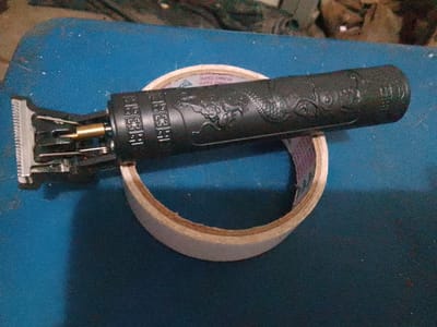 A close-up, eye-level shot focuses on a black cordless hair trimmer with an embossed dragon design, resting on a roll of light-colored tape. The trimmer is positioned horizontally, with its metallic blades and gold-colored shaft visible on the left. The tape roll, with visible "SUMO TAPE" text on its side, forms a circular base for the trimmer, which is angled slightly upwards. The background consists of a textured blue surface, likely a table or workbench, with some blurred, darker objects further in the background. The lighting is somewhat dim, creating shadows on the blue surface. The image quality is moderate, with some graininess. There are no people visible in the frame. The scene is not indicative of any specific location within the city of Cisarua, Indonesia.