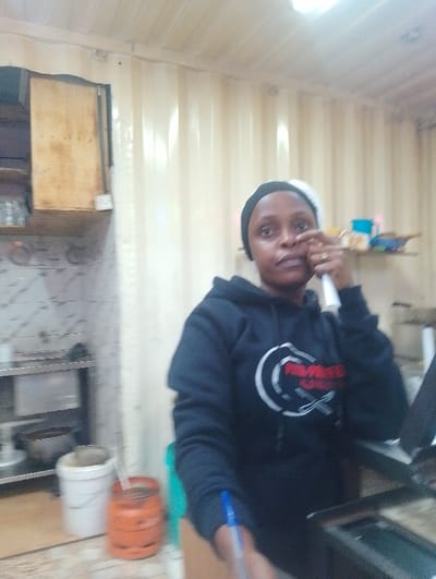 The image depicts a woman wearing a black hoodie with a red and white logo, standing in a kitchen located at coordinates -1.25 latitude, 36.684 longitude in Rungiri, Kenya. The woman has a white bandanna on her head. Her right hand is raised near her ear, holding a white object which may be a cell phone.  The room has a white corrugated metal ceiling and walls and an open doorway to the left. In the background, a stainless-steel counter with a metal shelf can be seen. To the right of the counter is a white plastic bucket, an orange propane tank, and a green object.  A black object on a table in the foreground appears to be a cash register.  A blue pen is on the table. The woman is the only person visible in the image.