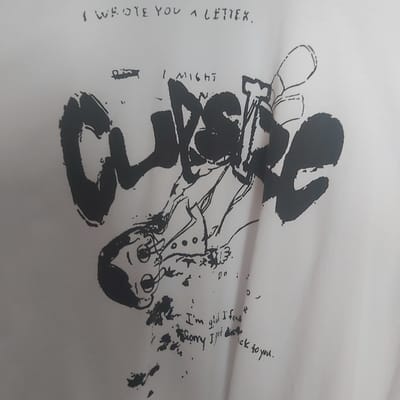 A white t-shirt displays a black graphic and handwritten text. The graphic features a cartoonish figure in the center, appearing to be falling or in distress, with large, stylized black lettering that reads "CURSE" partially overlaying it. Above the figure, a line of handwritten text reads "I WROTE YOU A LETTER," and below, smaller text states "I'm glad I found Sorry I got them back to you." The image is a close-up, focusing on the design and text. The location in Bobrov, Russia, is not discernible from the provided image.