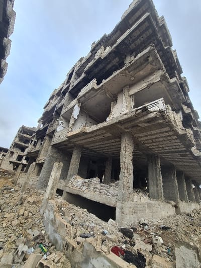 This image captures a severely damaged, multi-story concrete building within Joubar Municipality, Syria. The structure, appearing to be a former residential or commercial complex, exhibits extensive destruction. Numerous exterior and interior walls are absent, exposing multiple floor slabs and reinforced concrete support columns, many of which show fractured concrete and exposed rebar. The upper floors display significant dark discoloration and charring, indicative of fire or blast damage. The remaining concrete surfaces are pockmarked with numerous small impact marks, consistent with bullet or shrapnel strikes. The ground level around the building is covered in deep, irregular piles of concrete rubble and shattered building materials. Scattered fragments of miscellaneous debris, including colored plastics and fabric, are visible within the rubble in the foreground. A similarly damaged building is partially visible to the left. The sky is overcast and light grey. No individuals, actions, or interactions are present in the scene.