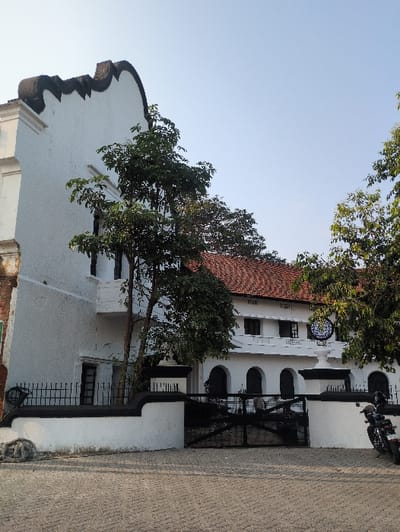 The image displays the exterior of the Pepper House, a colonial-era building situated in Fort Kochi, Kochi, India. The scene is illuminated by natural daylight under a clear, pale blue sky.
The main subject is a multi-story, predominantly white stucco building featuring a distinctive scrolled parapet painted black along its upper left edge. A reddish-brown tiled roof is visible above the lower white sections of the structure. The building's ground level is fronted by a low white wall with black decorative fencing, centrally interrupted by a large, two-part black metal gate. Above the gate, three arched doorways or windows are recessed into the white facade. To the right of these arches, a white circular emblem, resembling a clock face or stylized logo with a dark border, is mounted on the wall. A small white balcony extends from the upper left side of the building.
Several trees with green foliage are present, one standing directly in front of the left portion of the building, partially obscuring it. The foreground is paved with grey interlocking pavers arranged in a diagonal pattern. A loose coil of dark rope or cable rests on the ground near the base of the left wall. A black motorcycle, appearing to be a cruiser-style model, is parked on its kickstand to the far right of the frame. No individuals or active interactions are visible.