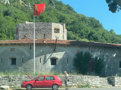 In Škaljari, Montenegro, a red Volkswagen Golf hatchback with its hood open is parked on the side of a paved road. The car is situated in front of a weathered stone building with a red-tiled roof. Above the building, a section of a stone fortress is visible, topped by a red flag with a yellow emblem flying from a flagpole. The fortress and building are set against a backdrop of lush green trees covering a steep hillside under a clear blue sky. A man wearing a light-colored shirt, shorts, a baseball cap, and sandals stands near the car, facing away from the viewer. He is carrying a dark bag slung across his shoulder. The scene suggests a pause in a journey, possibly due to a car issue, with a historic setting in the background.