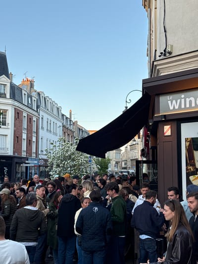 A bustling street scene in Reims, France, captures a large crowd of people gathered outside what appears to be a wine bar or restaurant. The image is taken from a slightly elevated perspective, looking down on the street and the individuals.

The foreground is dominated by the heads and torsos of numerous individuals, mostly adults, standing closely together. Many are looking towards the establishment, some appear to be socializing, while others seem to be waiting. Their attire suggests casual wear, suitable for an outdoor gathering.

On the right side of the image, a building features a commercial facade with a large display window showcasing bottles and other items, likely related to wine. Above this, a sign clearly reads "Le wine." Below the main sign, other partially visible text includes "Vins du monde" (Wines of the world) and "Le ba[r]". A red rectangular sign with "DH" and "IV" is also visible on the facade. A dark awning extends from the building over the outdoor area.

The background reveals a typical European street with multi-story buildings featuring varied architectural styles, including facades with brick and light-colored stone, and sloped roofs with dormer windows and chimneys. A few trees with white blossoms are interspersed among the buildings. The sky is clear and bright blue, indicating good weather, possibly late afternoon or early evening given the natural light and the illuminated shop sign.

Several street signs are visible: a blue sign with "JEAN JAURES" and a brown directional sign pointing towards "Hôtel le vergeur". On a building in the distance, a white sign with "À LOUER" (For Rent) can be seen. The overall impression is one of a lively urban environment, likely a popular spot for social gatherings.