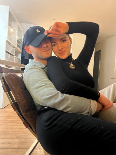A man and woman are seated intimately, facing the camera. The man wears a dark baseball cap and denim shirt. The woman, positioned above him, sports a black turtleneck and an ornate necklace. They appear to be sitting on a vintage-style chair with a brown leather seat. A white cabinet stands in the background, adding depth to the indoor setting. The image is rotated counter-clockwise.
