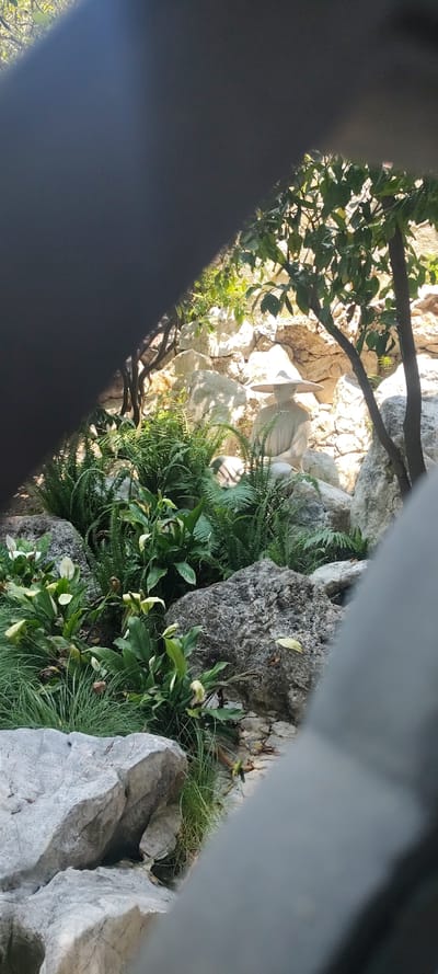 The image shows a partial view of a statue of a person with a wide-brimmed hat, situated among rocks, greenery, and trees. The statue is made of a light-colored material. The scene is partially obscured by a gray object in the foreground. The location is in Tetela Del Monte, Mexico.