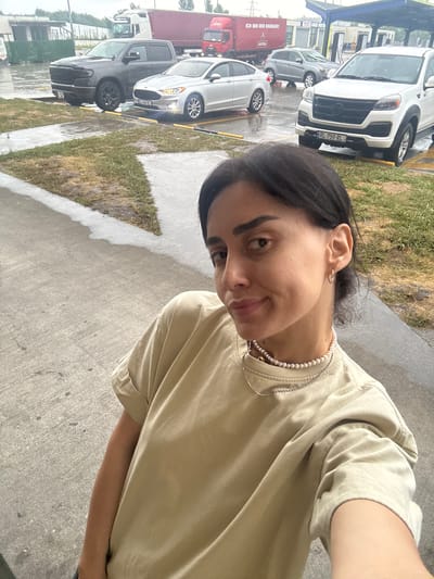 A medium shot captures a woman in a light brown t-shirt and two necklaces, a pearl strand and a silver chain, posing for a selfie outdoors. She has dark hair pulled back and is looking at the camera with a slight smile. Her left arm is visible, bent at the elbow. Behind her, a paved area, likely a parking lot, is wet from rain, with patches of grass and concrete. Several vehicles are parked in the background: a dark gray pickup truck, a silver sedan, a gray SUV, and a white SUV. A red truck with "ASTRA" written on its side is also visible. The location appears to be a service area with covered structures and charging stations. The overall environment suggests an overcast, possibly rainy day in a developed urban area. The woman is positioned in the foreground, with the vehicles and environment filling the background.