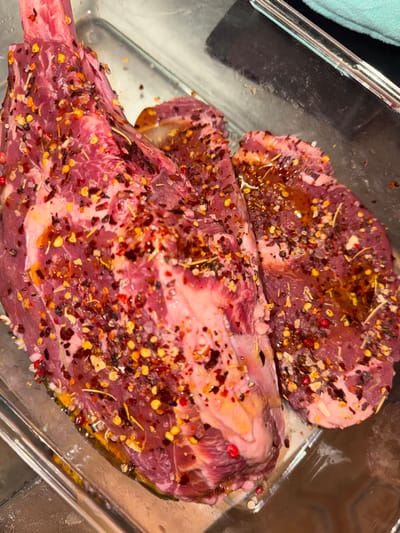 Two raw steaks marinating in a clear glass dish. The larger steak, with a bone, dominates the foreground. Both steaks are heavily seasoned with a colorful mix of spices, including red peppercorns, dried herbs, and other visible flakes. A thin layer of oil glistens on the surface of the meat. The dish's edges are visible, offering a clean contrast to the steaks.
