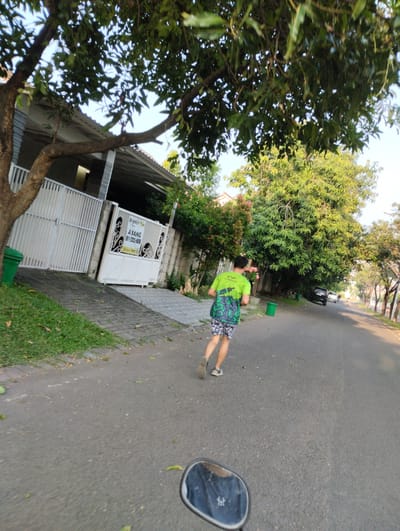 A person in a green t-shirt and patterned shorts is running on a paved road in the city of Sidodadi, Indonesia. The person is holding a bouquet of pink flowers. The road is lined with trees on the right side, and a sidewalk with a house and a white gate is on the left. A green trash can is visible on the left side of the road, and another on the right. The sky is clear and blue. The image is taken from a low angle, likely from a motorcycle.