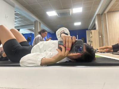 A person takes a mirror selfie with a smartphone. The individual wears a white button-down shirt and black shorts. A second individual stands in the background. The scene appears to be a gym or fitness center. The floor is tiled, and overhead lighting illuminates the room. The mirror reflects the person taking the photo and their surroundings, creating a mirrored perspective.