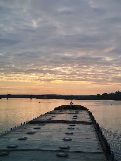 The image depicts a barge moving on a river under a sky with streaky clouds tinged with the orange light of sunset. In the distance, a bridge spans the river, and the opposite bank features trees and buildings. The camera's perspective is from the back of the barge, allowing a view of its large, flat surface with multiple hatch covers. The location is suggested to be the city of Нифантово, Russia.