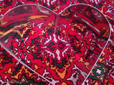 A heart-shaped cookie cutter, made of reflective metal, is placed on a red carpet with a complex geometric pattern. The carpet features intricate designs in red, yellow, green, and white. The cookie cutter's surface reflects the carpet's pattern, creating distorted and elongated shapes. The overall scene is an overhead shot, with the cookie cutter dominating the foreground and midground. The bottom left corner of the image displays the text "nubia Neo 3 5G". The specific location in Angren, Uzbekistan is not discernible from the image.