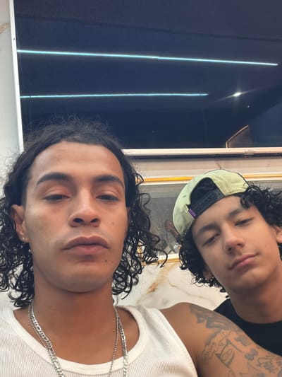 Two young males are captured in a selfie, with one in the foreground and the other slightly behind and to the right. The male in the foreground has long, dark, curly hair, brown eyes, and is wearing a white ribbed tank top and silver necklaces. He has a small silver earring in his left ear and visible tattoos on his right arm. The male behind him has shorter, dark, curly hair and is wearing a light green baseball cap backwards. He is wearing a black t-shirt. They appear to be indoors, with a reflective dark surface with horizontal light blue lines visible in the background, possibly a screen or a window. The overall lighting suggests an indoor setting, and the reflective surface could indicate a modern interior design. The image does not contain any discernible elements that would pinpoint a specific location within L'Hospitalet de Llobregat, Spain.