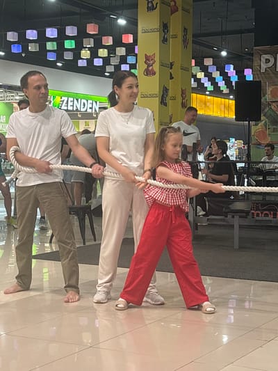 In what appears to be an indoor retail or entertainment space in Tri Protoka, Russia, three individuals are engaged in a tug-of-war activity with a thick rope. A man on the left, barefoot, wears a white t-shirt and khaki pants. Beside him, a woman also in a white t-shirt and light-colored pants, wearing sneakers, holds the rope. On the right, a young girl in a red and white checkered top and red pants, wearing sandals, actively participates, pulling the rope with focused effort. In the background, other shoppers and a speaker system are visible. The environment features a high ceiling with colorful, cube-shaped lights and decorative pillars with cat imagery.