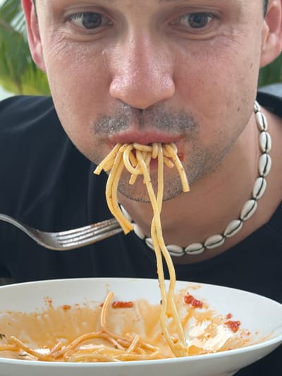 The media file captures a close-up, low-angle shot of a person eating spaghetti. The person, whose face is partially visible, is wearing a necklace made of white shells. They are in the process of lifting a forkful of spaghetti to their mouth. The spaghetti is coated in a sauce, and some sauce and pasta remnants are visible on a white plate to the right of the frame. The background is dark, suggesting an indoor setting, possibly at night. The lighting is soft, highlighting the texture of the person's skin and the food. The scene appears to be taking place in Baan Bang Rak, Thailand, although no specific environmental cues are visible in the frame to confirm this location. No text is visible in the image.