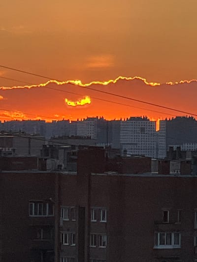 The image shows a city skyline at sunset, with the sun peeking through the clouds and casting an orange glow. In the foreground, there are several brick buildings with visible windows. Behind them, a line of taller, more modern buildings can be seen. The sky is a vibrant orange and yellow, with darker clouds obscuring some of the light. The scene appears to be a typical urban landscape, with a focus on the interplay of natural light and man-made structures. The location is Ladozhskoe Ozero, Russia, which is a city on the eastern shore of Lake Ladoga.