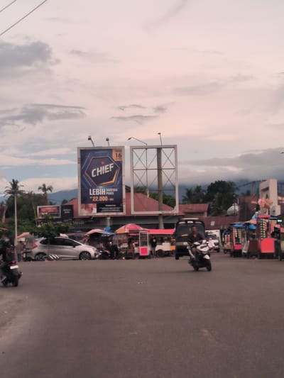 This medium shot captures a bustling street scene in Seulimeum, Indonesia, under an overcast sky. The diffused light suggests either early morning or late afternoon, or simply a heavily cloudy day.

In the foreground, a wide asphalt road occupies the bottom half of the frame. Several motorcycles are visible: one in the far left foreground, another with a rider in the center-right, and a third partially visible in the middle ground near a silver car. People are also present, some appearing as motorcyclists, others standing near stalls or vehicles.

The middle ground features a variety of activity. A silver car is parked on the left side of the road, partially obscured by a small tree. Numerous colorful street food stalls and vendors line both sides of the street, suggesting a market area. Buildings with red-tiled roofs are visible behind the stalls. In the background, lush green trees and distant mountains shrouded in haze complete the scenic view.

Dominating the upper half of the image are two large billboards. The one on the left prominently displays a product advertisement with a large hexagonal logo and the word "CHIEF." Text beneath it reads "LEBIH BANYAK PUAS" (More satisfaction) and "RP 22.000/BKS" (Rp 22,000/pack). Smaller text includes "NEW 16 BATANG" (New 16 sticks/cigarettes) and "PERINGATAN:" (Warning:). Another smaller billboard to the left reads "WARUM SUPER KRETEY" and "DATACO BARU," with "SAFARI" also visible. The billboard on the right appears to be an empty frame or under construction. A phone number, "08216507 0047," is also visible on a structure in the mid-right.