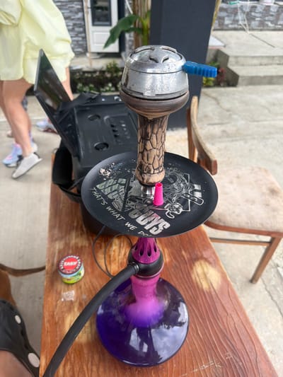 This media file depicts a close-up, rotated view of a hookah setup on a wooden table. The main subjects are the hookah components: a metallic base with intricate carvings, a wooden stem, a black disc with white graphics, a purple hose, and a violet glass base containing a purple liquid. A small, blue, ridged piece, likely a poker or valve, is visible on top of the metallic base. A small, pink object, possibly a grommet, is attached to the stem. A small, circular tin, possibly containing tobacco or accessories, is also on the table.

The scene appears to be outdoors, with hints of a paved ground and possibly a building or wall in the background. A chair with a wooden frame and what appears to be a fabric seat is partially visible behind the hookah. The lighting suggests it might be daytime, with some shadows indicating natural light. There are no people prominently featured in the shot, though a sliver of a person's arm and legs, wearing light-colored clothing and white sneakers, is visible at the bottom left, suggesting someone is either setting up or using the hookah.

The black disc on the hookah stem features graphics and text. The text visible reads "UNS THAT'S WHAT WE DO". The overall impression is a relaxed, informal setting, possibly a backyard or patio, where someone is preparing to use the hookah. The location is identified as Baranovka, Russia.