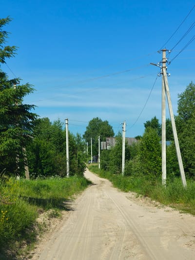 The image shows a dirt road curving through a rural area, flanked by trees and vegetation. Several utility poles with power lines are visible along the road. The sky is clear and blue. There are no people or animals visible in the image. The scene appears to be a quiet, sunlit day in the countryside, possibly on the outskirts of the city of Zhukovo, Russia, given the presence of traditional Russian rural elements like utility poles and simple buildings. The road shows tire tracks, indicating it is used by vehicles. Some low-lying plants and wildflowers are visible along the sides of the road.