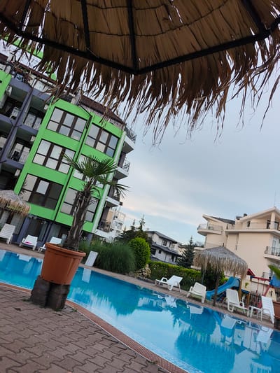 The image depicts a resort swimming pool area in the city of Ravda, Bulgaria. A large, brightly lit green building with multiple balconies and windows dominates the left side of the frame. To the right, a lighter-colored building with modern architectural elements is visible. The foreground is occupied by a bright blue swimming pool, with its surface reflecting the surrounding structures and sky. Around the pool, several white lounge chairs are arranged on a brick-paved patio. A large, brown planter with a palm tree stands prominently near the pool's edge. Overhead, a thatched umbrella partially obscures the view, its dry fronds hanging down. The sky above is overcast. In the background, lush green foliage and other buildings can be observed. A children's play area with a blue slide and a red structure is also present near the pool.