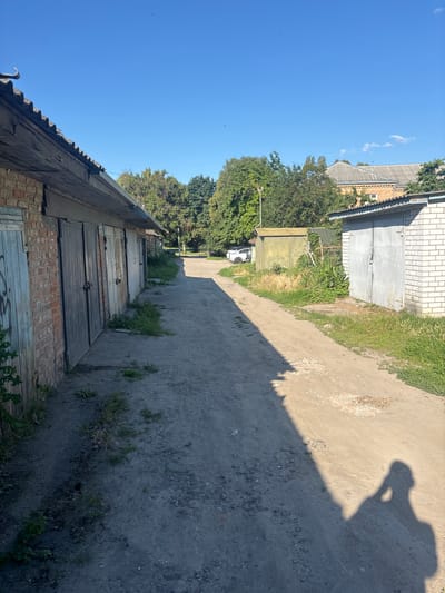 A person's shadow is cast on a dirt path lined with brick and metal garages. The garages have closed doors and some are covered in overgrown vegetation. A white car is parked in the distance, near a row of trees. The sky is clear and blue. This scene is located in the city of Pereiaslav, Ukraine.