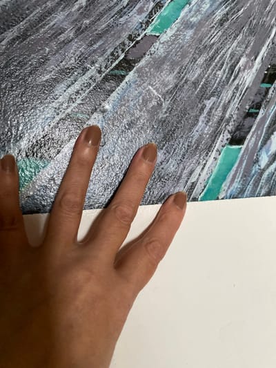 The image shows a close-up of a hand with light brown skin and light brown nail polish, resting on a textured surface with streaks of gray, black, white, and teal. The hand is positioned in the lower left and center of the frame, with four fingers and a portion of the thumb visible. The fingers are extended and gently pressed against the textured surface, which appears to be a painting or a textured wallpaper. The background is a stark white surface, likely a wall or a table, occupying the bottom portion of the image. The image is taken from a slightly elevated angle, looking down at the hand and the textured surface. There is no indication of the specific location within Bílina, Czechia.