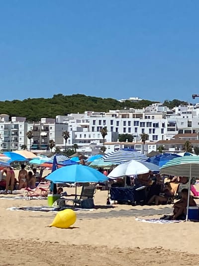 The image depicts a crowded beach scene in Conil de la Frontera, Spain. In the foreground, a yellow inflatable object lies on the sand. The midground is filled with numerous beach umbrellas providing shade for beachgoers, some of whom are seated on chairs or towels. Beach tents and trolleys are also visible. In the background, white buildings with multiple stories are clustered together, backed by a dense green hillside. The sky is clear and blue.