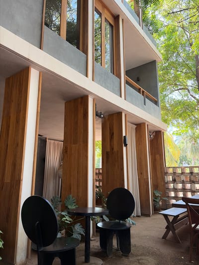 This media file captures a modern, multi-story building from a low angle, looking upwards towards lush green tree canopies and a bright sky. The architecture combines grey concrete, light beige walls, and warm wooden accents, including window frames and balcony structures.
The building features multiple levels, each with what appears to be a private balcony or terrace. These outdoor spaces are finished with wooden flooring and low, white railings. White fabric shades or curtains are drawn on several balconies, providing privacy or sun protection. Small ceiling fans are visible attached to the underside of the balconies, suggesting a warm climate. Large windows reflect the surrounding trees, integrating the natural environment with the modern structure.
On a lower level or ground area to the right, there are black, minimalist outdoor tables and chairs set on a sandy or dirt surface. A potted plant with large, green leaves, possibly a Monstera, sits beside the furniture. A wooden slatted fence or screen is also visible on the far right.
The scene depicts a tranquil, outdoor setting, likely a resort or residential building in a tropical location, consistent with Brisas de Zicatela, Mexico. The time of day appears to be bright daylight, with clear, sunny weather. No people or visible text are present in the image.