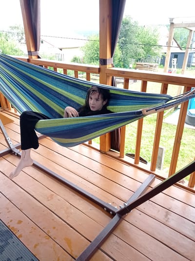 A young person with dark hair is lying in a striped hammock on a wooden deck. They are wearing black pants and a black top. Their left leg is extended out of the hammock, and their bare foot is visible, with a small tattoo on the ankle. The hammock has blue, green, and yellow stripes and is supported by a metal stand. The deck appears to be part of a porch or balcony, with wooden railings and support beams. In the background, there are trees, houses, and a cloudy sky. The setting suggests an outdoor recreational area, possibly a backyard or a campsite. The lighting indicates it is daytime, with overcast weather. The person's facial expression is neutral, suggesting relaxation or contemplation. No visible text is present in the image. The location context is Degtyarsk, Russia.