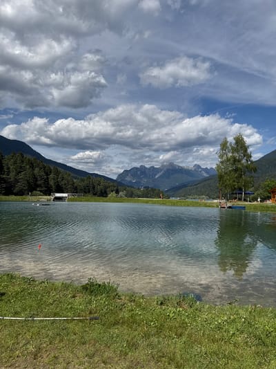 The image depicts a serene lakeside scene, likely in Calalzo di Cadore, Italy. The perspective is from the edge of a body of water, looking towards a tree-lined shore with imposing mountains in the background. The sky is a brilliant blue, adorned with dramatic, fluffy white clouds that cast shadows, suggesting it is daytime, possibly late morning or early afternoon.
The water's surface is rippled, reflecting the sky and the surrounding greenery. A sandy bottom is visible in the shallow areas near the shore. On the right side of the frame, a lush green grassy bank slopes gently towards the water. A long, white object, possibly a fishing rod or a surveying pole, lies horizontally on the grass, near the water's edge. Further out in the water, a small red marker float is visible.
The left side of the image features a dense forest covering the lower slopes of the mountains. The peaks of the mountains are jagged and rocky, reaching towards the sky. A tall tree with green foliage stands prominently on the shore, casting a shadow. In the distance, a small structure or building can be seen on the shoreline, along with what appears to be a pier or platform extending into the water. A few indistinct figures are visible near the shore and on the platform, engaged in leisure activities, though their specific actions are not discernible. The overall impression is one of a peaceful, idyllic natural setting, bathed in sunlight and offering opportunities for recreation. There is no visible text in the image.