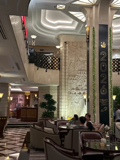 The image shows an indoor scene in what appears to be a hotel lobby or a fine dining establishment in Shanghai, China. The scene is decorated for a New Year's celebration, likely for the year 2020, as indicated by a prominent banner displaying "2020" in large gold lettering, along with the text "Happy New Year." A graphic of a horse is also visible on the banner.
Several people are present, primarily seated in lounge chairs and at tables in what seems to be a seating area. They appear to be engaged in conversation and relaxation. The decor is elegant, with ornate architectural features, including curved ceilings and decorative moldings. A large, detailed bas-relief sculpture depicting historical figures or a historical event is mounted on the wall, adding to the sophisticated ambiance.
The lighting suggests it is evening or indoors with artificial lighting, creating a warm and inviting atmosphere. There are various decorative elements such as plants, lamps, and framed pictures on the walls, contributing to the luxurious setting. The overall impression is one of a festive and upscale gathering.