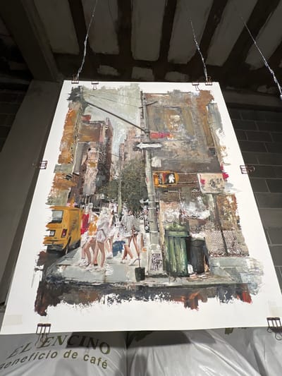 A large, unframed, vertically oriented artwork depicting a textured, impressionistic urban street scene is suspended in an industrial or storage environment. The artwork portrays a city street corner with several multi-story buildings, a partial view of a yellow van with a partially visible license plate ("H377") on the left, and a group of approximately five to seven pedestrians walking across a crosswalk or sidewalk in the foreground. A street pole featuring a pedestrian traffic signal displaying a white "walk" symbol and an illegible street sign is central on the right. Below the signal, two dark green waste bins are positioned near a chain-link fence. Overhead, utility lines crisscross the upper portion of the painted cityscape.
The artwork is secured by four metal binder clips attached to its top and bottom edges, from which thin wires extend upwards, suspending it from an unfinished concrete slab ceiling with exposed joists. To the left of the artwork, a raw concrete column is visible, while a gray cinder block wall is present to the right. Directly beneath the artwork, several large white bags are stacked, with text "EL ENCINO" and "beneficio de café" clearly visible on one of them. This suggests the setting is a coffee processing or storage facility located in the city of León, Mexico.