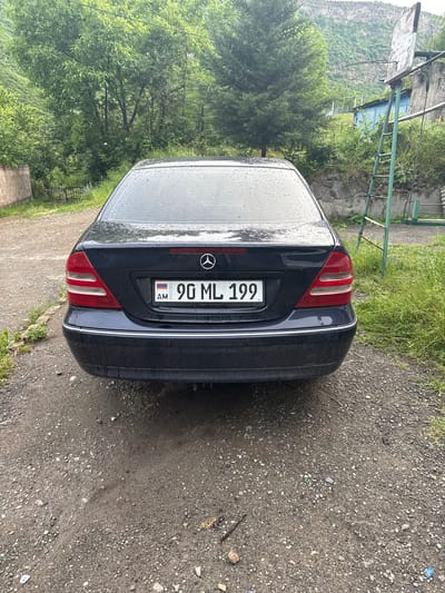 A dark blue Mercedes-Benz sedan is parked on a gravelly, uneven surface in front of lush green trees and a mountain in the background. The car's license plate reads "AM 90 ML 199". To the right of the car, a weathered wooden basketball backboard is mounted on a green metal pole with a ladder attached. The area appears to be a residential or semi-rural setting, possibly in the city of Tumanyan, Armenia, given the context. The sky is overcast, and raindrops are visible on the car's rear window and trunk.