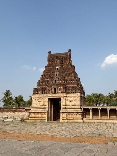The image shows the Hazara Rama Temple in Kamalapura, India, situated under a bright blue sky with scattered clouds. The temple's structure is primarily constructed of tan stone, exhibiting layered details and architectural designs. A pathway with stone pavers leads towards the structure, with adjacent vegetation in the surrounding area. The building's orientation in the frame shows a partial view of the entrance and its prominent tower.