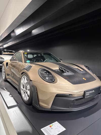 The image shows a gold-colored Porsche 911 GT3 RS on display at the Porsche Museum in Stuttgart, Germany. The car is positioned at an angle, with its front facing towards the right of the frame. It features a carbon fiber hood with prominent vents, large alloy wheels, and racing decals on its side. The car is situated on a black display platform. In the background, the museum's architecture is visible, characterized by dark, angled ceiling elements and spotlights. A white Porsche is partially visible behind the gold car. A sign reading "PORSCHE MUSEUM" is mounted on the front of the gold car. A printed document with energy efficiency ratings is placed on the floor in front of the car. The lighting in the museum is ambient and focused on the displayed vehicles.