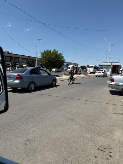 The image depicts a street scene in Pakhtakor, Uzbekistan, under a clear blue sky. In the foreground, a light blue Chevrolet sedan is parked on the right side of the road. Further down the street, a person is riding a bicycle. Several people are gathered near a white bus and a small building with a blue "Ucell" sign. In the background, other vehicles, including a white truck and a silver sedan, are visible. The environment appears to be an urban area with buildings, trees, and streetlights lining the road. Power lines crisscross the sky.