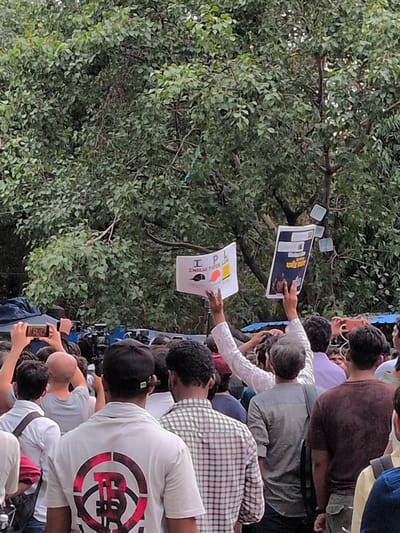 This image captures a crowd of people gathered outdoors, likely at a protest or rally, with lush green trees forming a backdrop. The orientation of the image is vertical, and it appears to be daytime. Several individuals are holding up signs, suggesting a demonstration or public gathering. One sign, held by a person in the center, displays the text "I P INDIA" in large, bold letters, with a red heart symbol and a mustache graphic. Another sign, visible in the upper left portion of the frame, has a dark blue background with white text and images, though the text is not clearly legible.
The people in the image are dressed in casual clothing, with a variety of tops and headwear visible. Some individuals are looking towards the signs, while others are engaged with their phones, possibly taking pictures or videos of the event. The atmosphere seems active and engaged, with people close together in a relatively dense crowd. The presence of trees and natural foliage suggests an outdoor setting, possibly a park or a street lined with greenery. No specific time of day is explicitly indicated, but the lighting suggests daylight. The overall impression is one of a public assembly with a focus on visible messaging through signs.