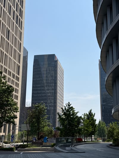 The image shows a view of the Chaoyang District in China, framed by modern architecture. The sky is clear and blue. In the foreground and background, parts of buildings with repeated rectangular patterns are visible. A taller, darker glass building stands in the middle ground. Trees and greenery line a sidewalk and street to the right. A sign is visible advertising "luckin coffee". The image appears to be taken from a slightly low angle looking upward.