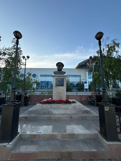 The image depicts a bust of a man in military attire, likely a memorial, situated outdoors in front of a modern building. The bust is mounted on a stone pedestal which is adorned with red and white flowers. The pedestal itself is located on a tiered stone platform. Two ornate lampposts flank the platform, and a black metal fence is visible in the foreground. The background shows a building with a blue roof and large windows. Trees and a clear sky with some wispy clouds complete the scene. The memorial is located in Yakutsk, Russia, and is dedicated to Hero of the Soviet Union Vladimir Ivanovich Kuzmin, who was born and raised in Yakutia and died in battle during the Great Patriotic War.