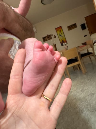 A baby's foot is being held in an adult's hand, which has a gold ring on the ring finger. The baby is wearing a diaper. The background shows a room with furniture and artwork on the wall. The location is not discernible from the image.
